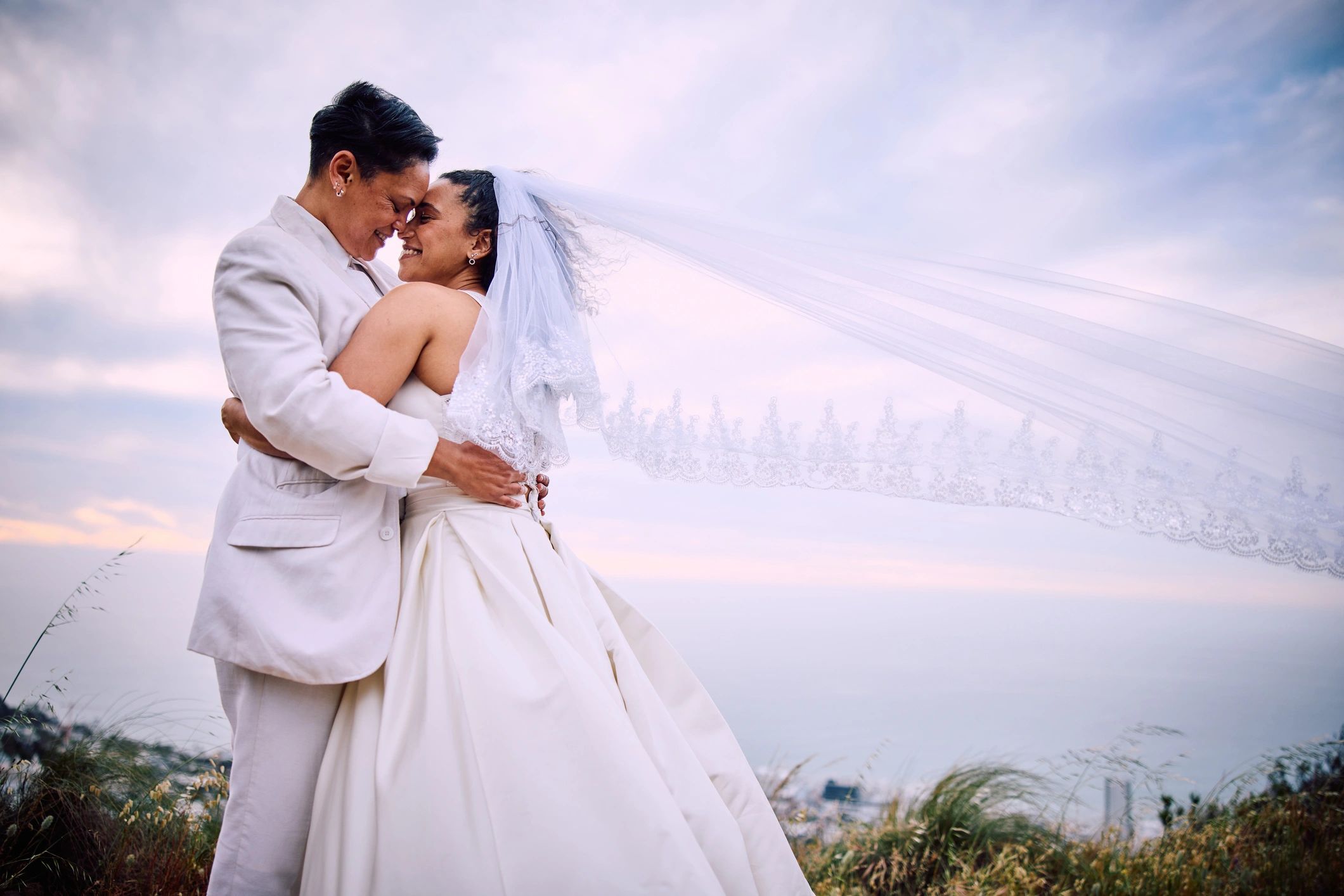 Newlywed couple sharing a romantic kiss beneath a bridal veil outdoors at sunset